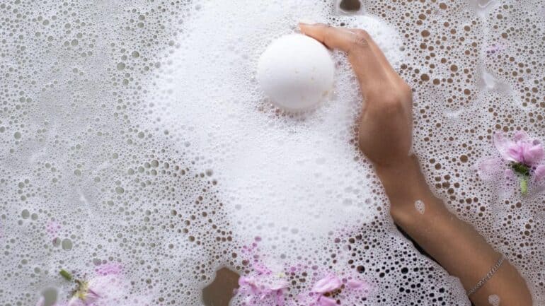 Woman's hand holding bath bomb in bath with petals.