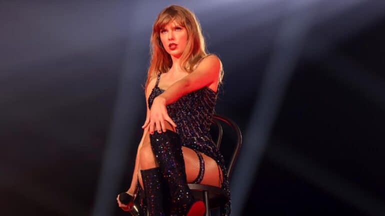 Taylor Swift in black costume sitting on a chair on stage.