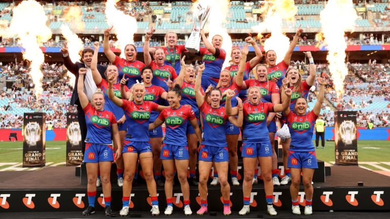 NRLW Knights team holding up the trophy on stage with fire behind them.
